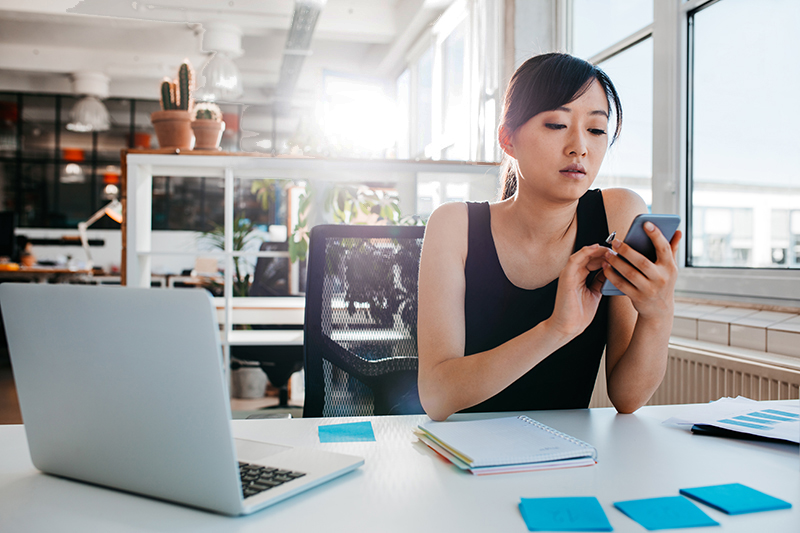 Portrait of young Asian woman sitting at a desk with laptop and using mobile phone. Asian businesswoman working in modern office. Requesting a prescription refill or other service such as motion sickness prevention, COVID treatment with Paxlovid, urinary tract infection, bladder infection, UTI, yeast infection treatment.