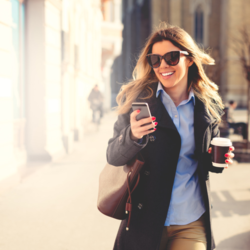 Young smiled woman looking at smart phone and holding cup of coffee. She is walking at city square.
